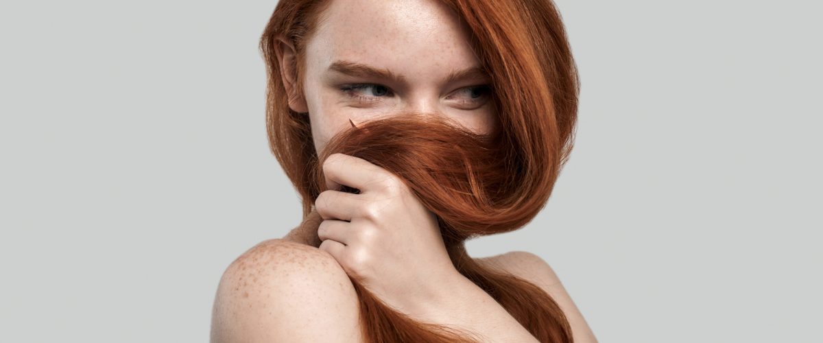 Studio shot of young and gorgeous redhead woman playing with her long hair while standing against grey background. Beauty portrait